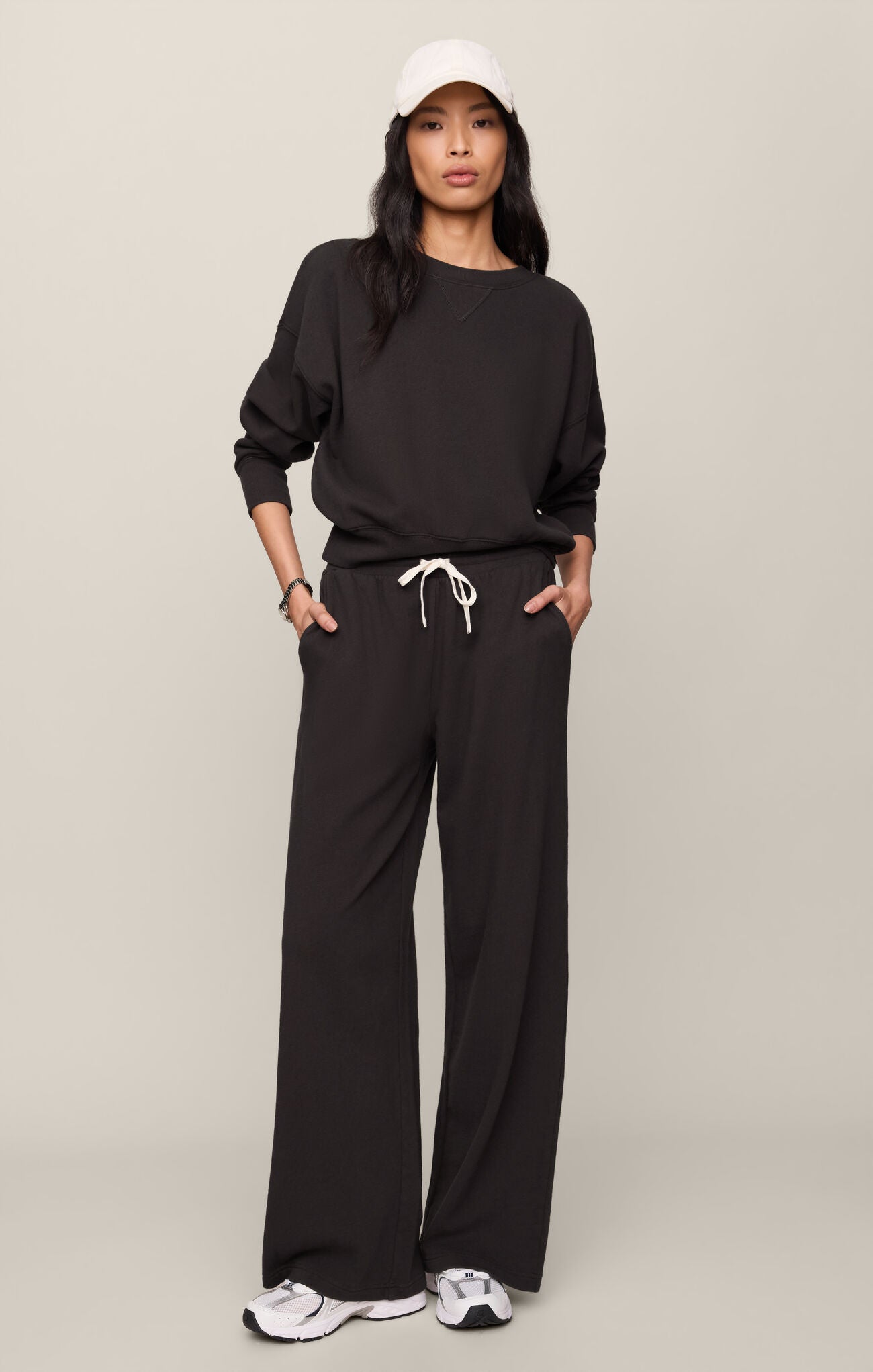 Person wearing a black outfit with wide-leg pants and a white cap on a beige background
