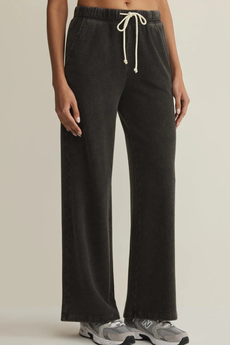 A person wearing dark charcoal wide-leg sweatpants with a cream drawstring, paired with gray sneakers, shown from the waist down against a plain light beige studio background.