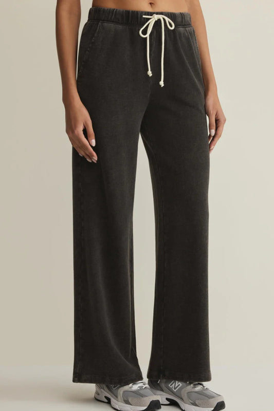 A person wearing dark charcoal wide-leg sweatpants with a cream drawstring, paired with gray sneakers, shown from the waist down against a plain light beige studio background.