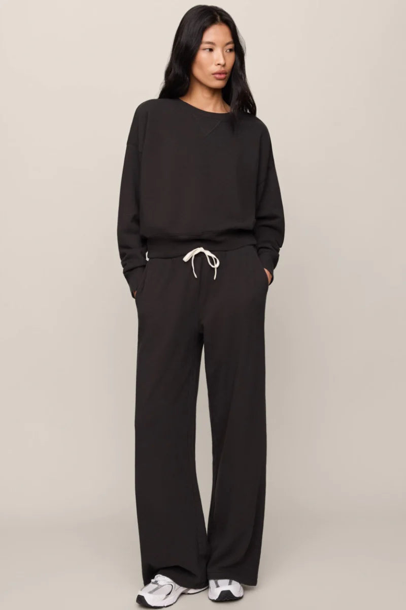 A woman modeling a black two-piece lounge set consisting of a long-sleeve sweatshirt and matching wide-leg sweatpants, featuring a white drawstring at the waist, against a plain beige studio background.