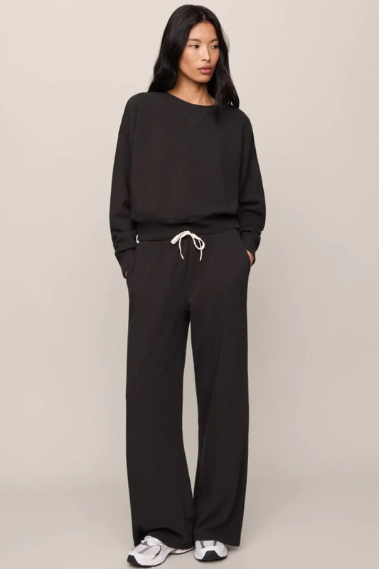 A woman modeling a black two-piece lounge set consisting of a long-sleeve sweatshirt and matching wide-leg sweatpants, featuring a white drawstring at the waist, against a plain beige studio background.